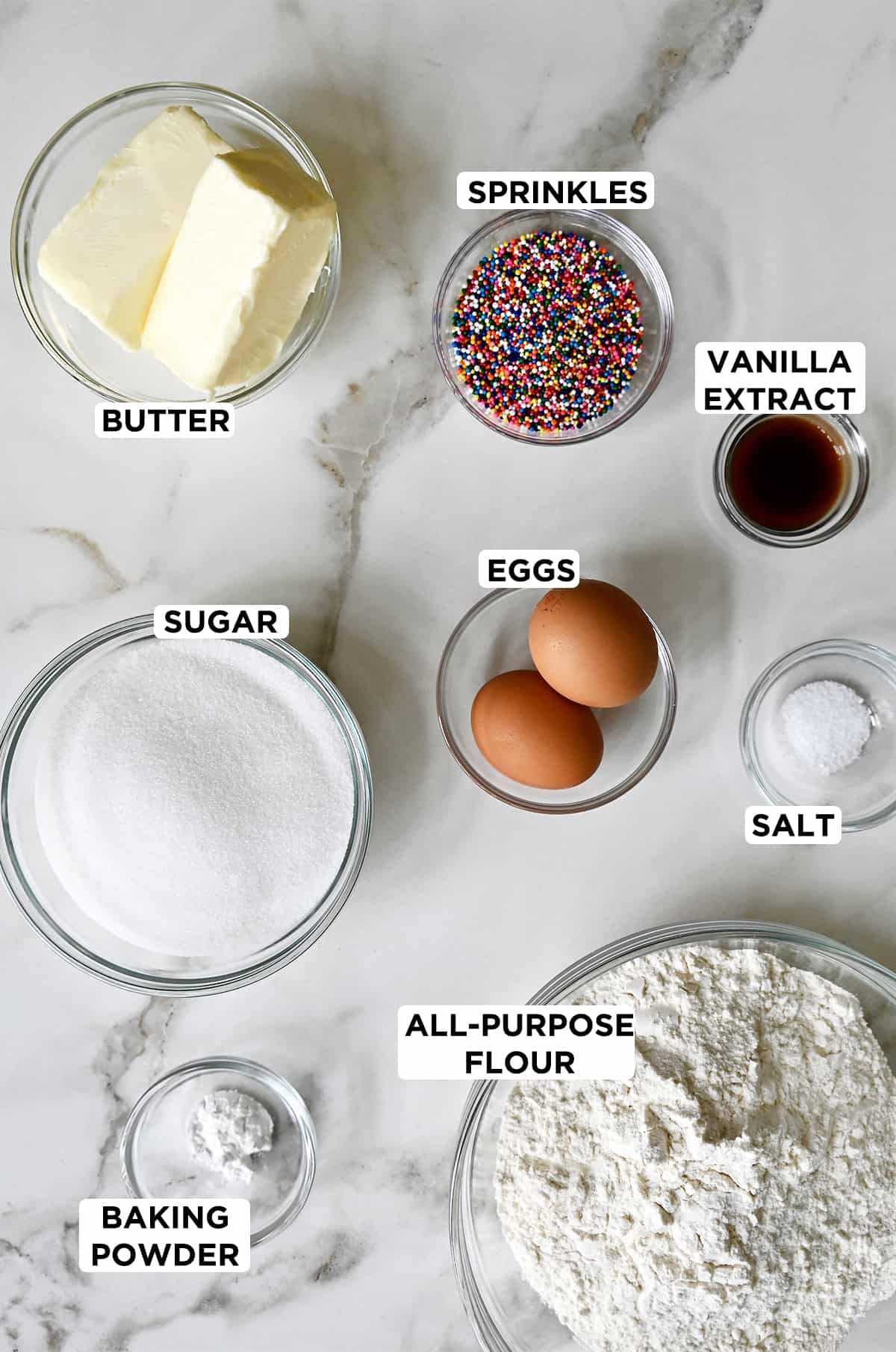 Various sizes of clear bowls containing ingredients to make butter cookies, including rainbow sprinkles, vanilla extract, two eggs, salt, all-purpose flour, baking powder, white sugar and butter.