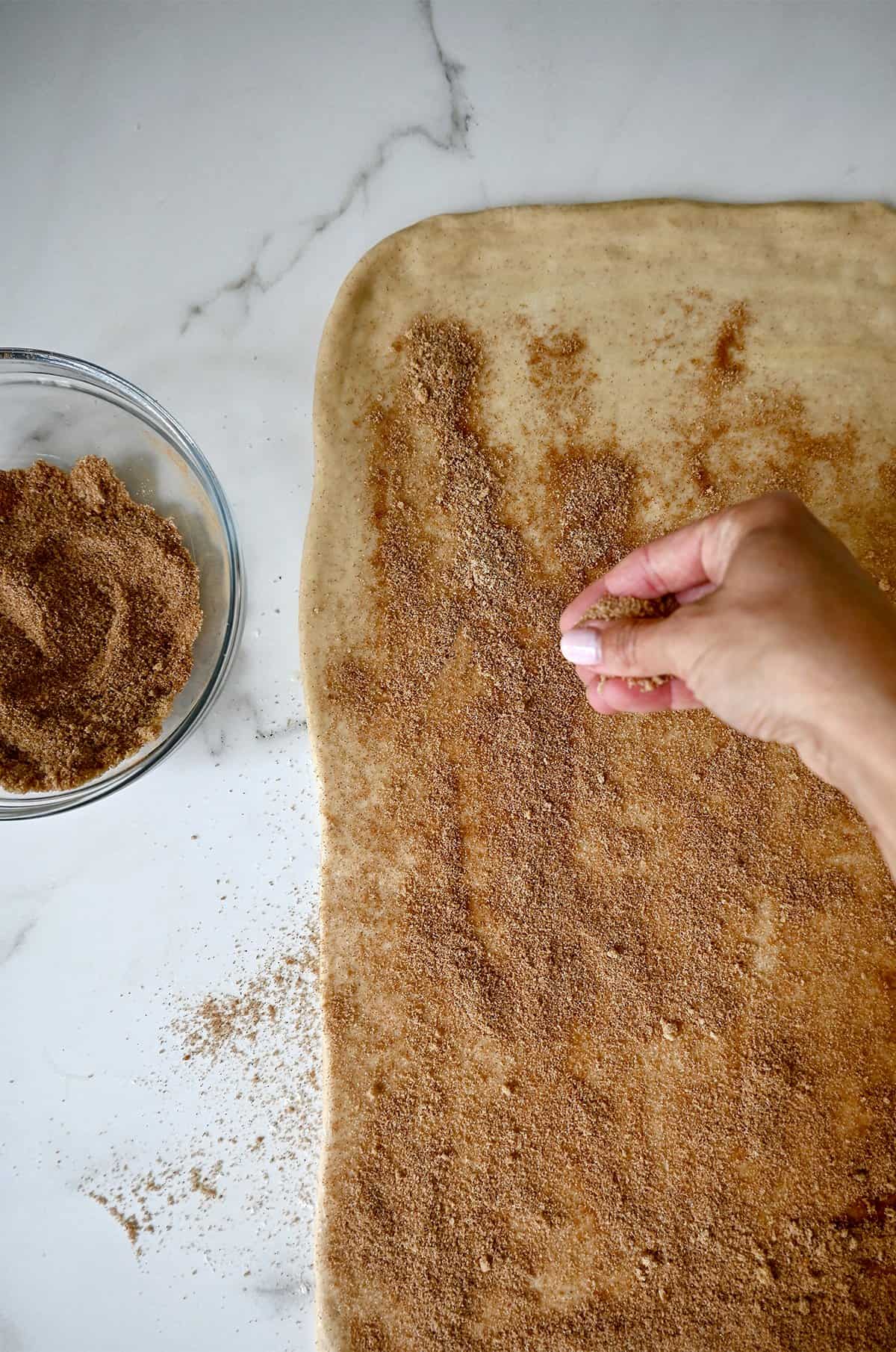 Sprinkling cinnamon sugar on top of cinnamon roll dough that's been brushed with melted butter.