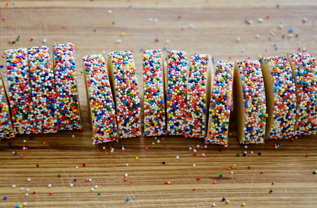 A log of cookie dough with rainbow sprinkles cut into slices on a wood cutting board.