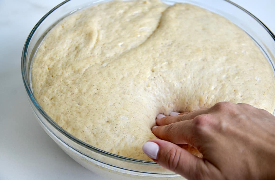 A hand pressing down sticky bun dough in a clear bowl