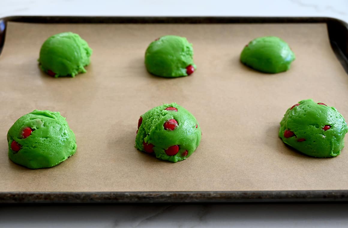 Scoops of unbaked green-colored cookie dough with red M&Ms on a parchment paper-lined baking sheet