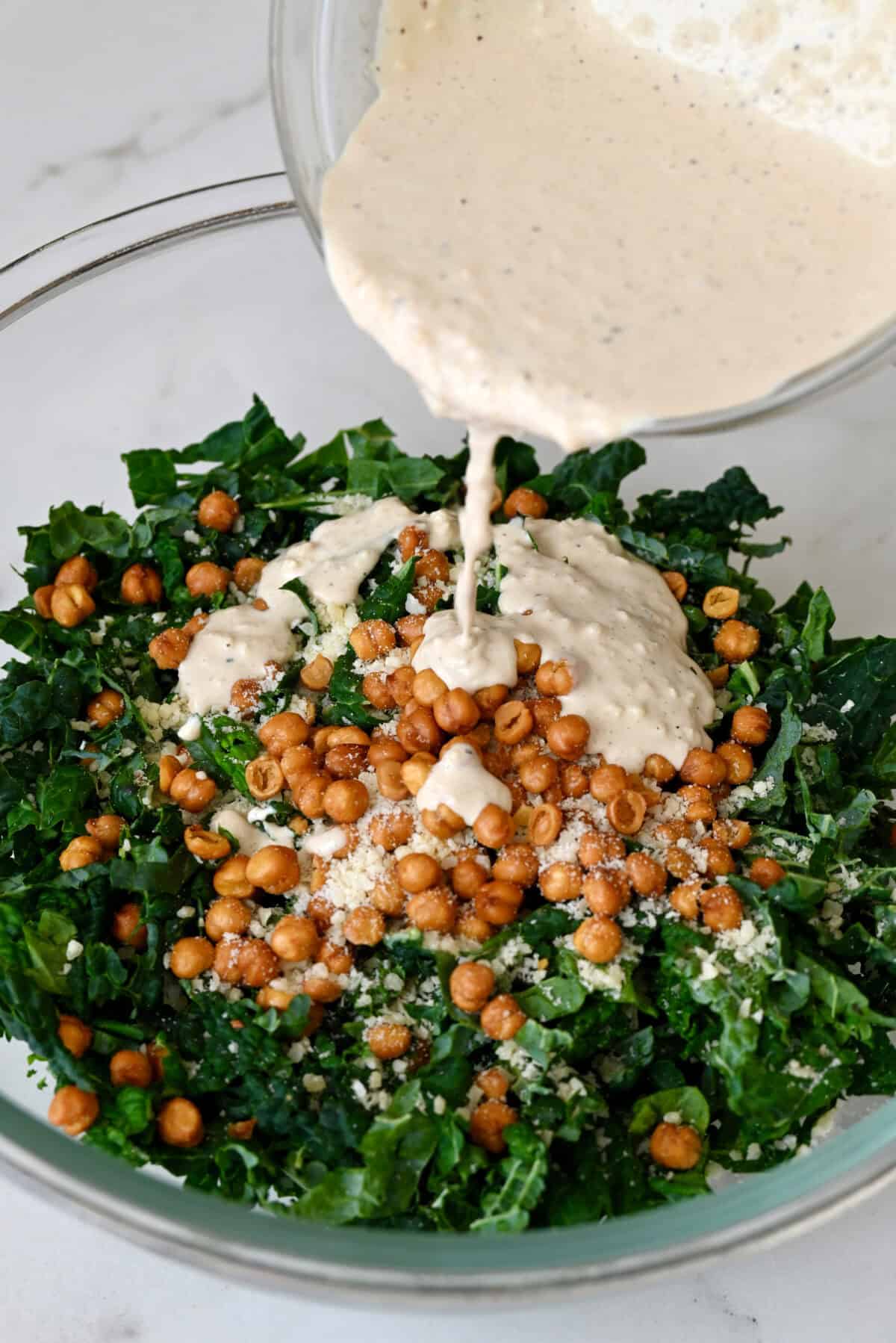 Adding homemade Caesar dressing to a bowl containing shredded kale and crispy chickpeas.