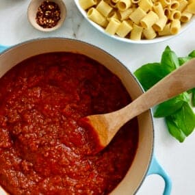 A large stock pot containing Secret Ingredient Bolognese Sauce and a wooden spoon next to a large bowl containing rigatoni noodles