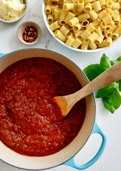 A large stock pot containing Secret Ingredient Bolognese Sauce and a wooden spoon next to a large bowl containing rigatoni noodles