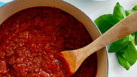 A large stock pot containing Secret Ingredient Bolognese Sauce and a wooden spoon next to a large bowl containing rigatoni noodles