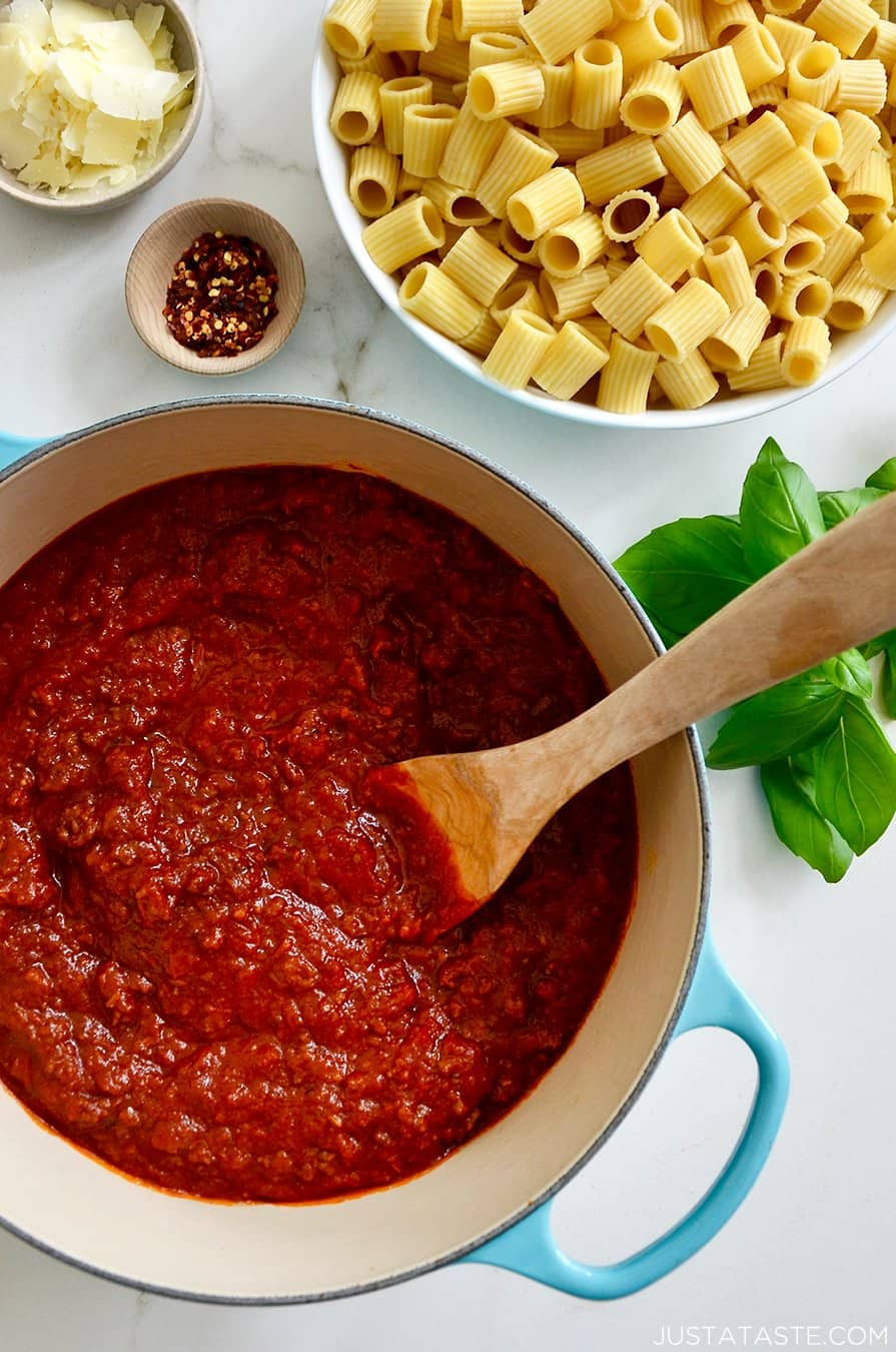 A large stock pot containing Secret Ingredient Bolognese Sauce and a wooden spoon next to a large bowl containing rigatoni noodles