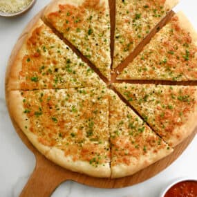 A top-down view of Cheesy Garlic Bread Pizza slices atop a wood serving board surrounded by small bowls containing marinara sauce, parmesan and fresh herbs