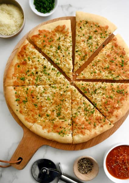 A top-down view of Cheesy Garlic Bread Pizza slices atop a wood serving board surrounded by small bowls containing marinara sauce, parmesan and fresh herbs