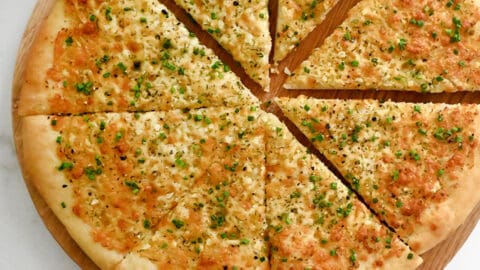 A top-down view of Cheesy Garlic Bread Pizza slices atop a wood serving board surrounded by small bowls containing marinara sauce, parmesan and fresh herbs