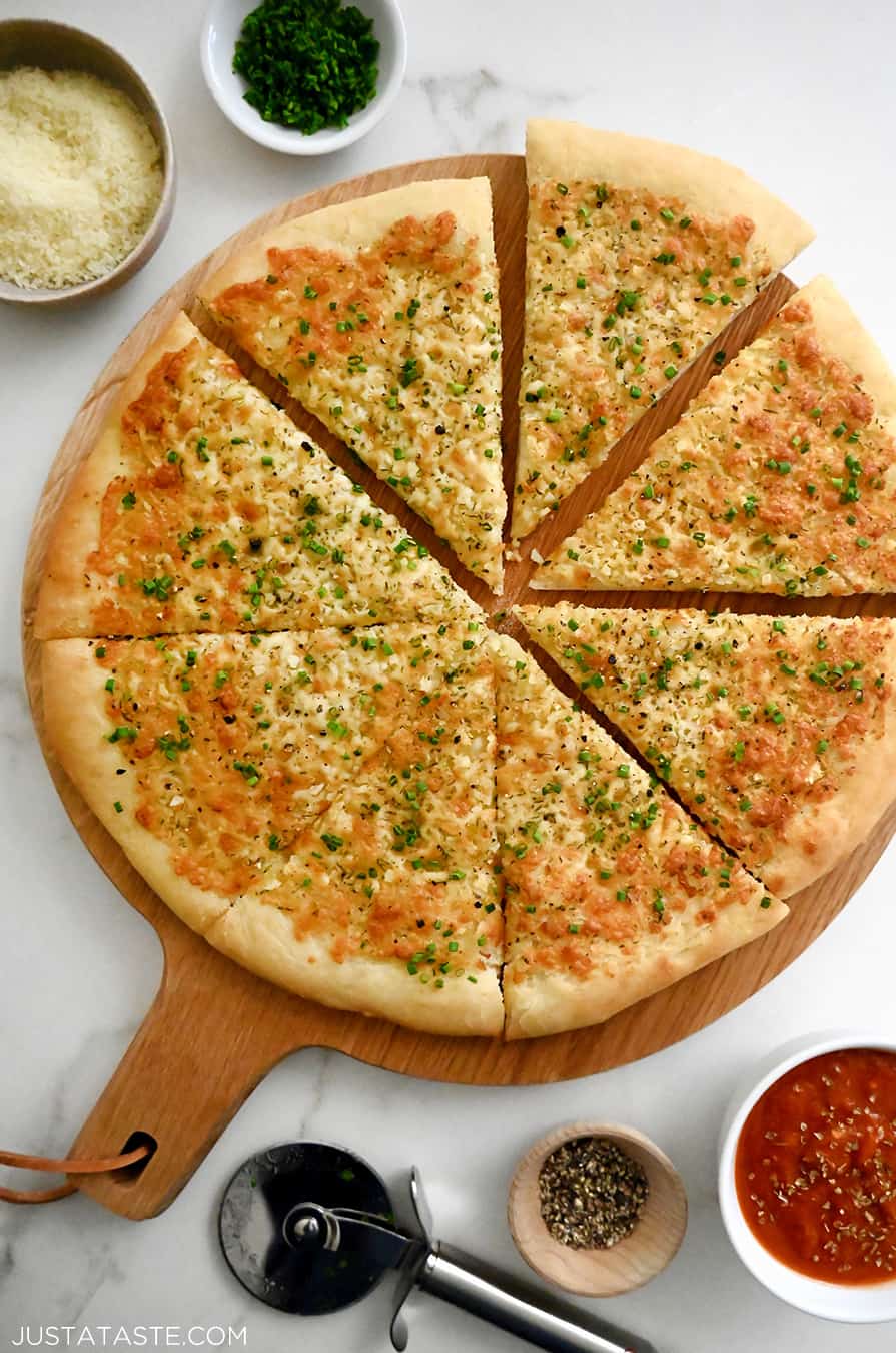 A top-down view of Cheesy Garlic Bread Pizza slices atop a wood serving board surrounded by small bowls containing marinara sauce, parmesan and fresh herbs