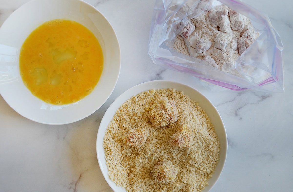 Bowls containing beaten eggs and breadcrumbs and a bag with chicken nuggets in flour.
