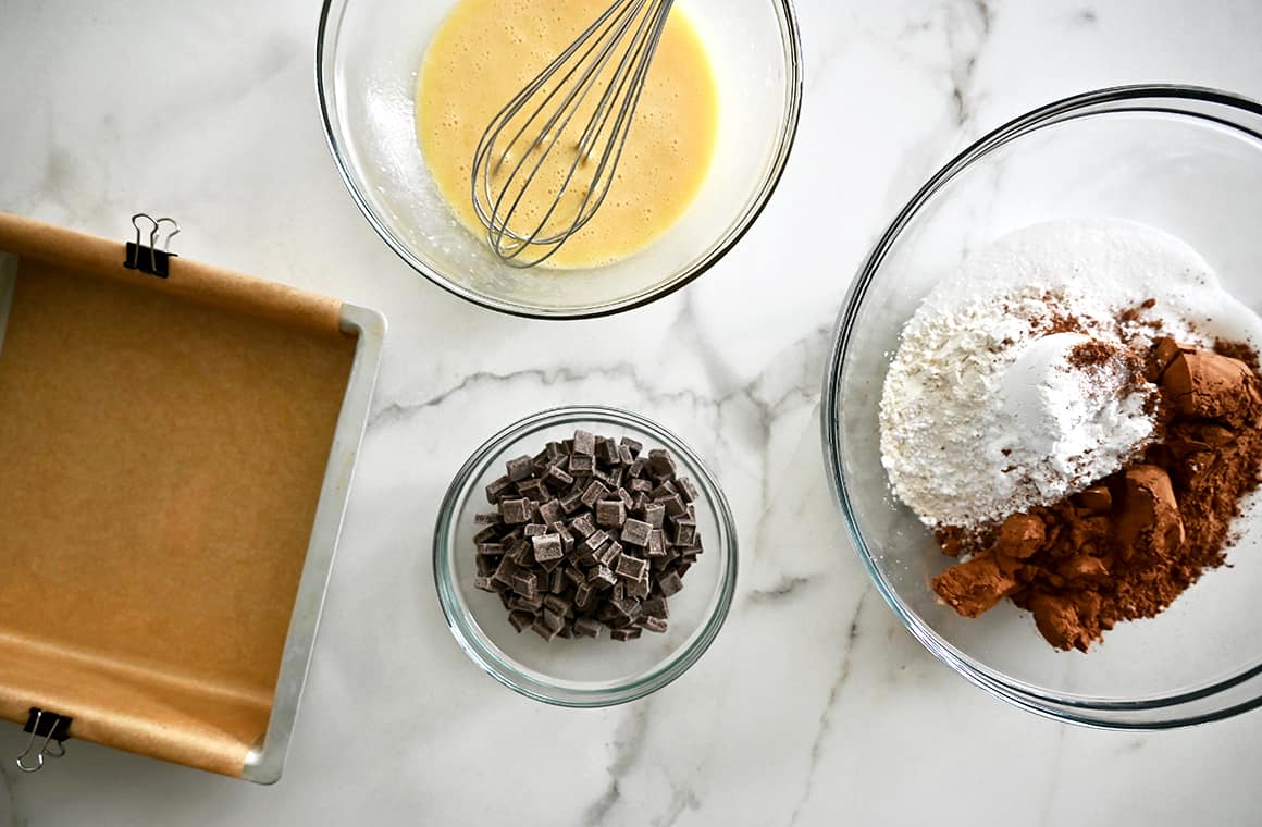 A top-down view of a parchment paper-lined baking dish, a clear bowl containing whisked eggs with a whisk, a small clear bowl with chocolate chunks, and a clear bowl with flour and cocoa powder