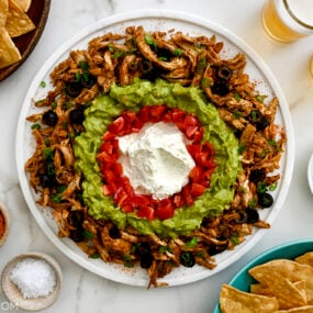 A white platter with taco dip surrounded by bowls of chips and seasonings