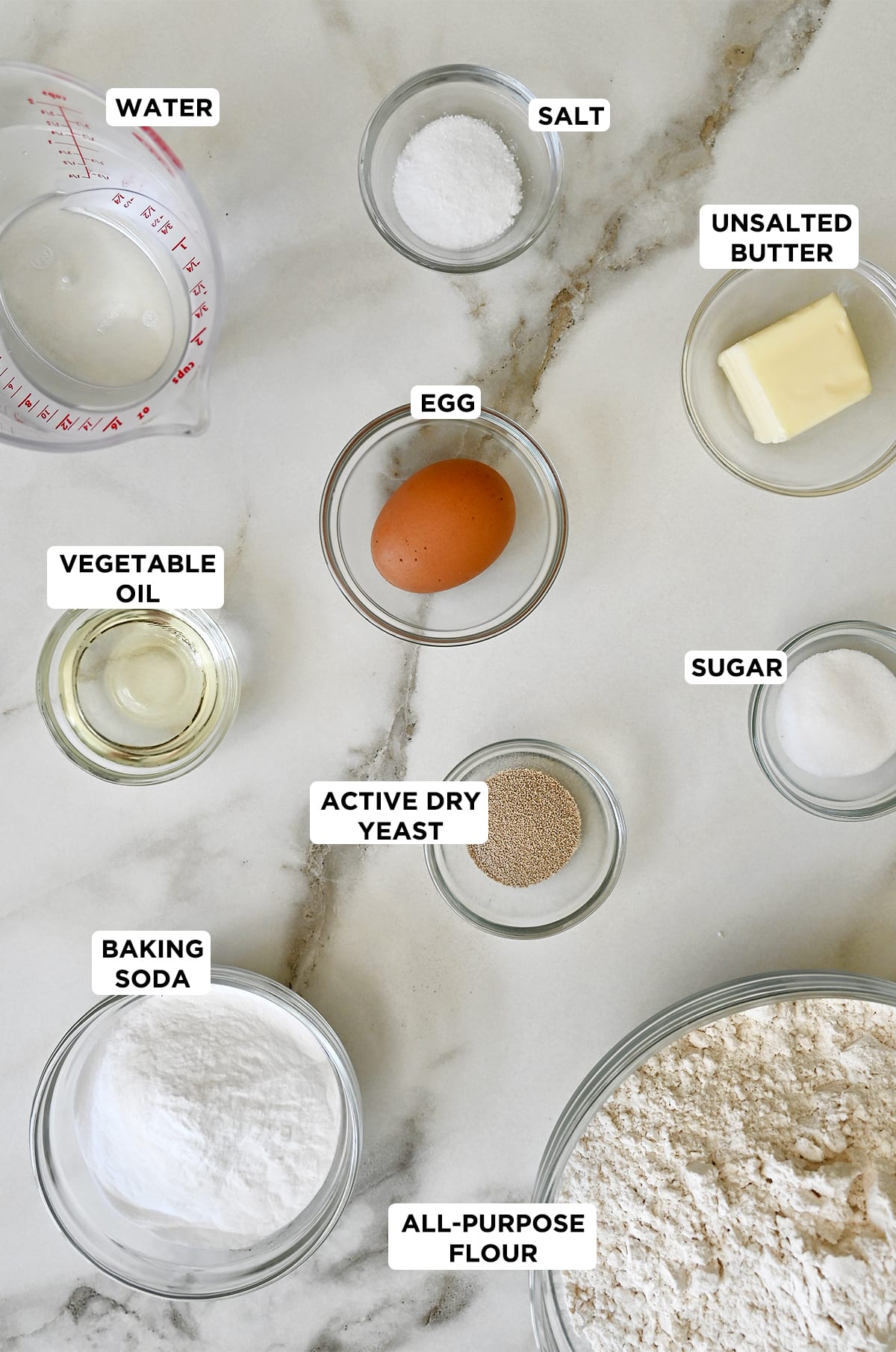 Various sizes of glass bowls containing ingredients to make soft pretzels, including water, salt, butter, sugar, active dry yeast, all-purpose flour, baking soda and vegetable oil. 