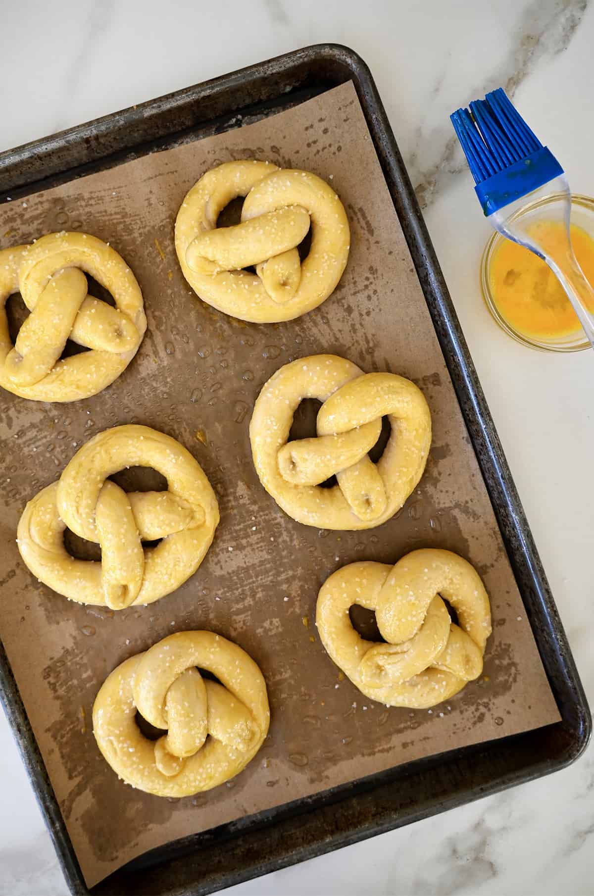 Six unbaked pretzels on a baking sheet topped with an egg wash and sprinkled with salt. A small bowl containing egg wash and a pastry brush are next it.