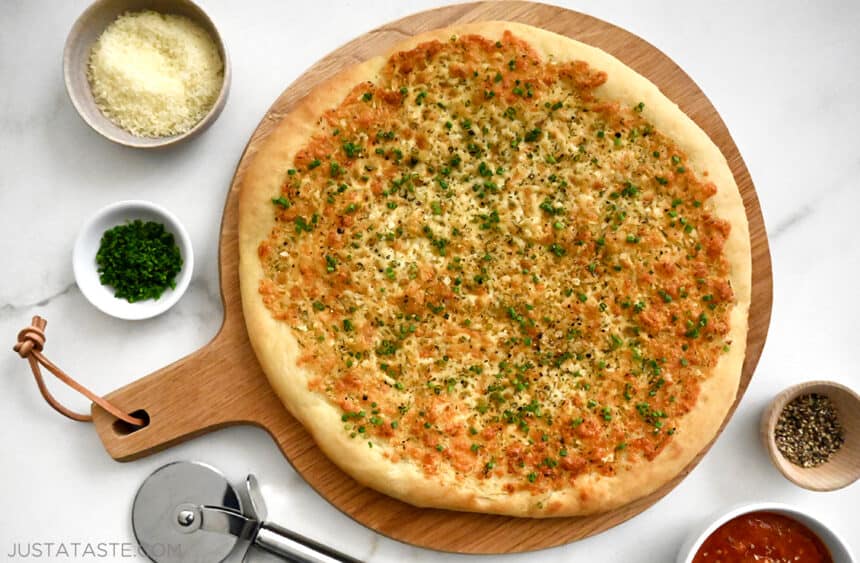 The best Cheesy Garlic Bread Pizza on a wood serving board next to a pizza cutter and small bowls containing parmesan cheese, fresh herbs and black pepper