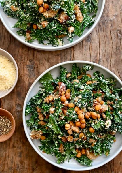 Two plates containing kale salad tossed with homemade Caesar dressing, topped with croutons and crispy chickpeas.