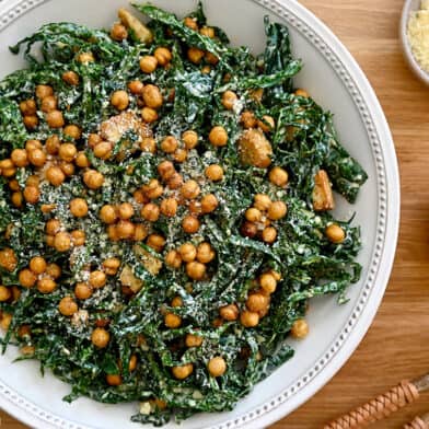 A top-down view of kale salad in a white bowl with lemons and a small bowl of Parmesan cheese on the side