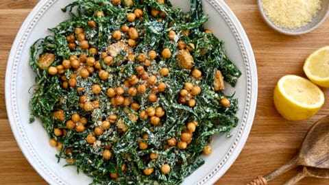 A top-down view of kale salad in a white bowl with lemons and a small bowl of Parmesan cheese on the side