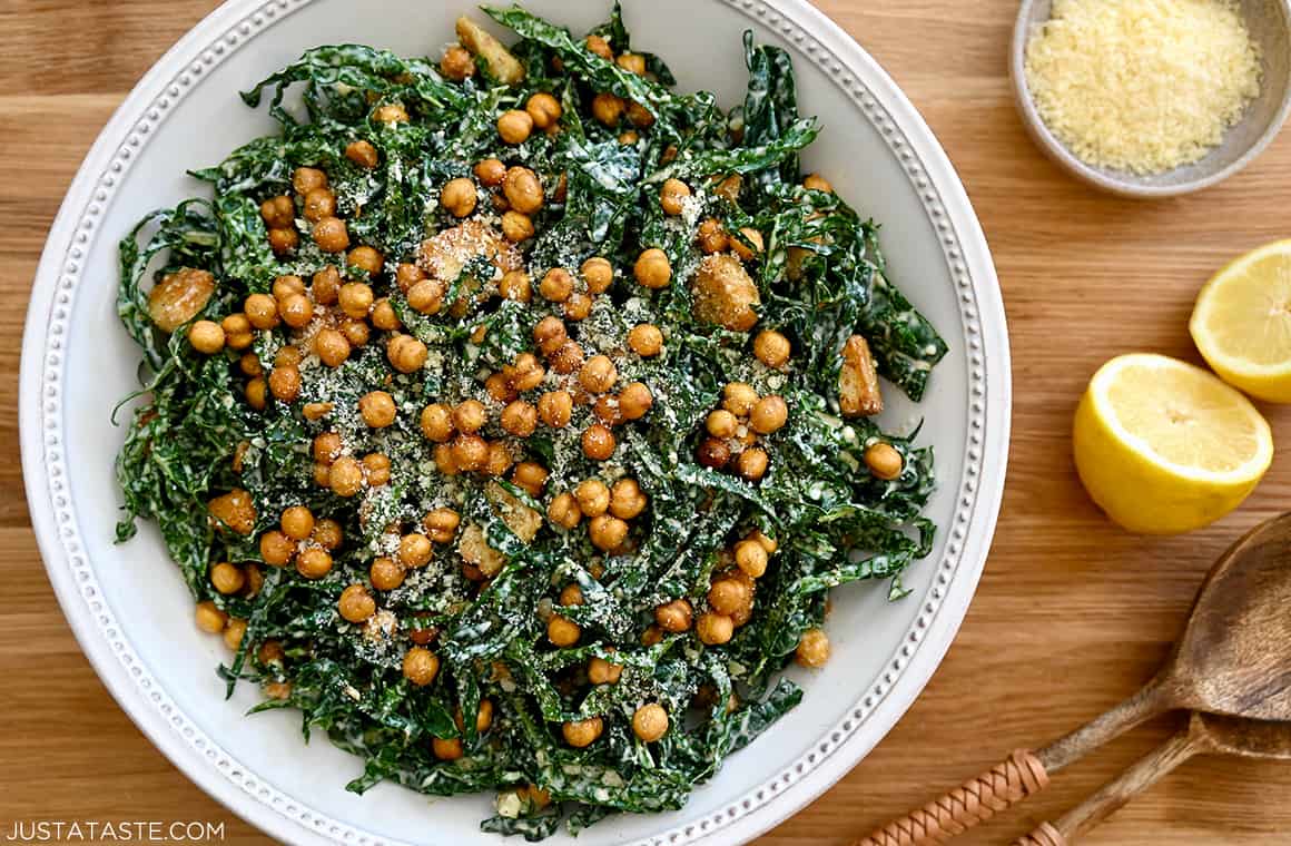 A top-down view of kale salad in a white bowl with lemons and a small bowl of Parmesan cheese on the side