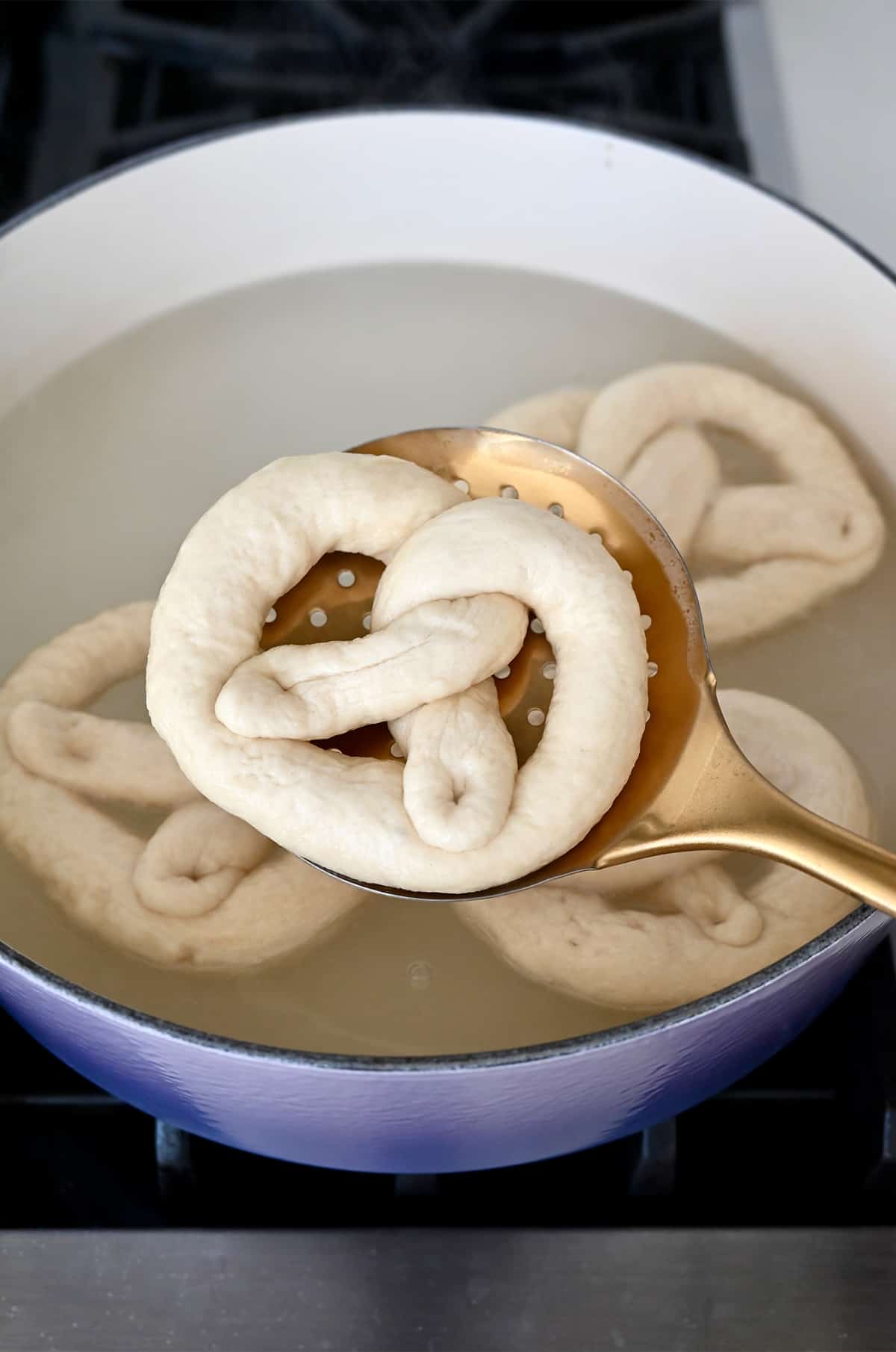 A slotted spoon with an unbaked pretzel over a Dutch oven containing a baking soda bath.