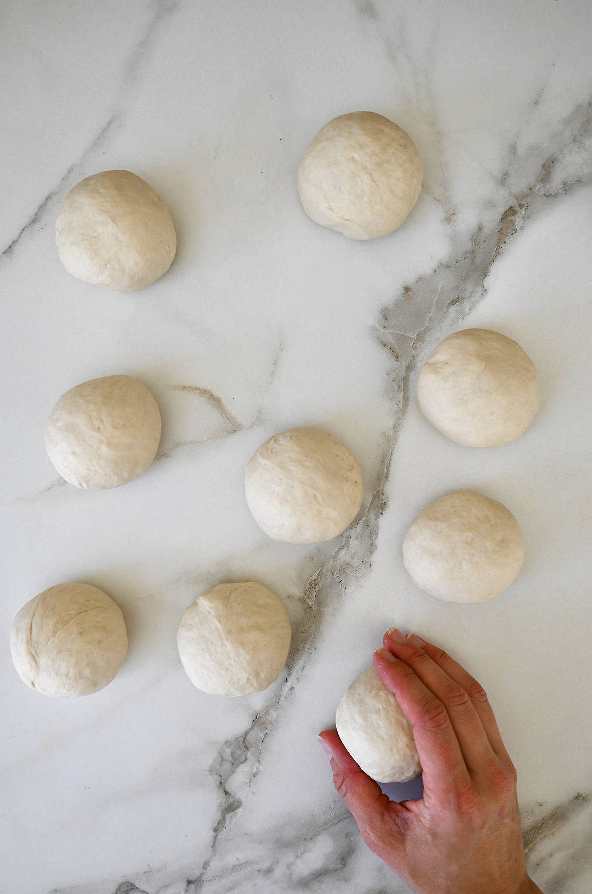 Nine dough balls on a marble surface with a hand holding onto one of them.