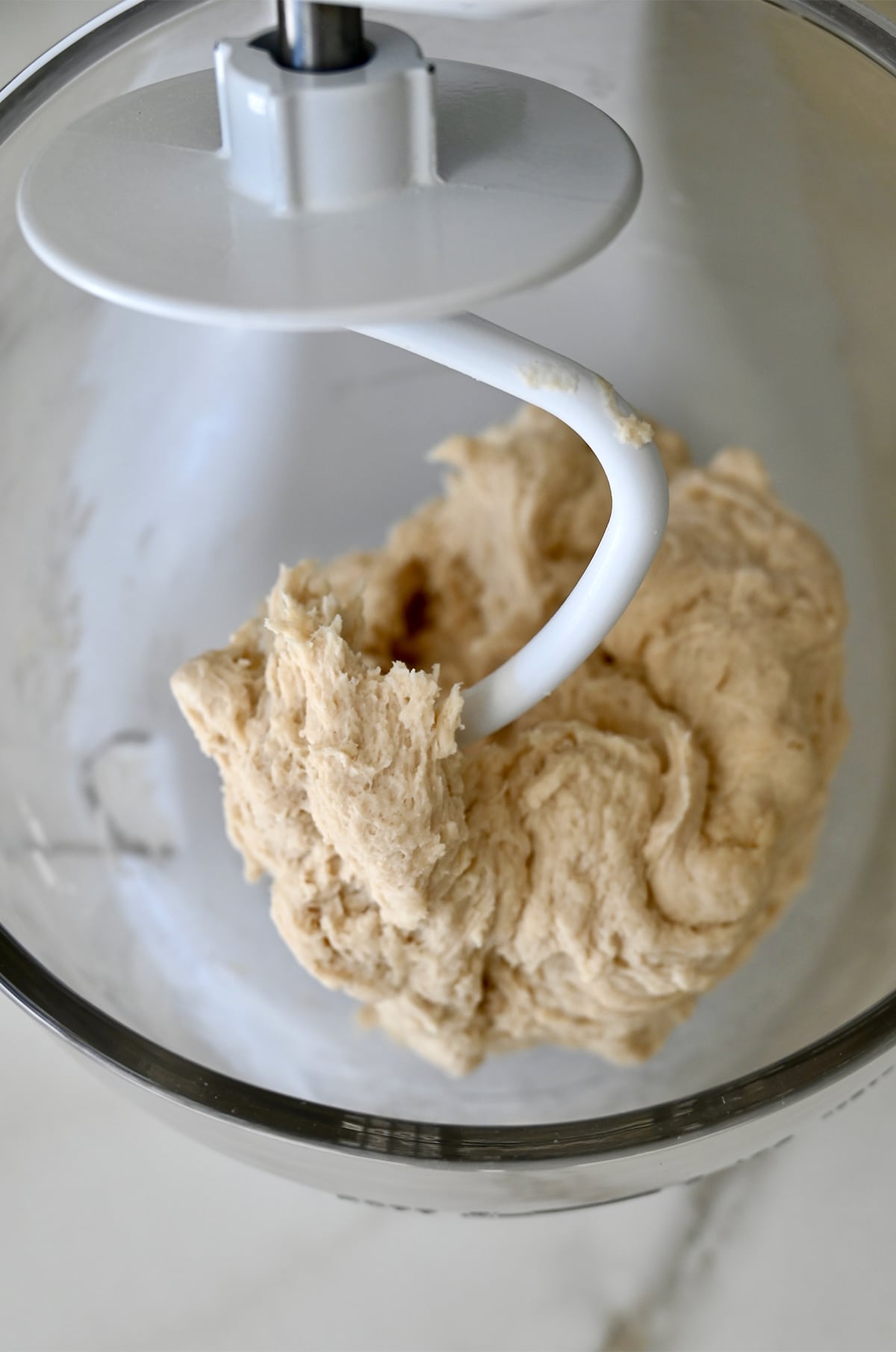 Pretzel dough wrapped around the dough hook of a white stand mixer with a glass bowl.
