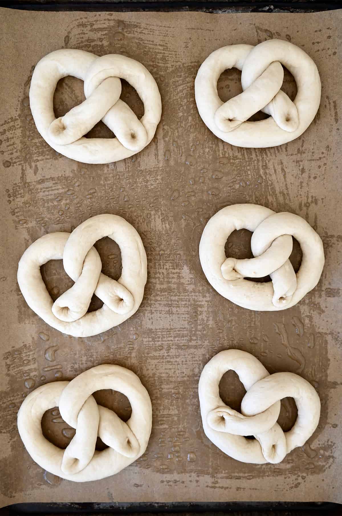 Six iconically-shaped unbaked pretzels on a parchment paper-lined baking sheet.