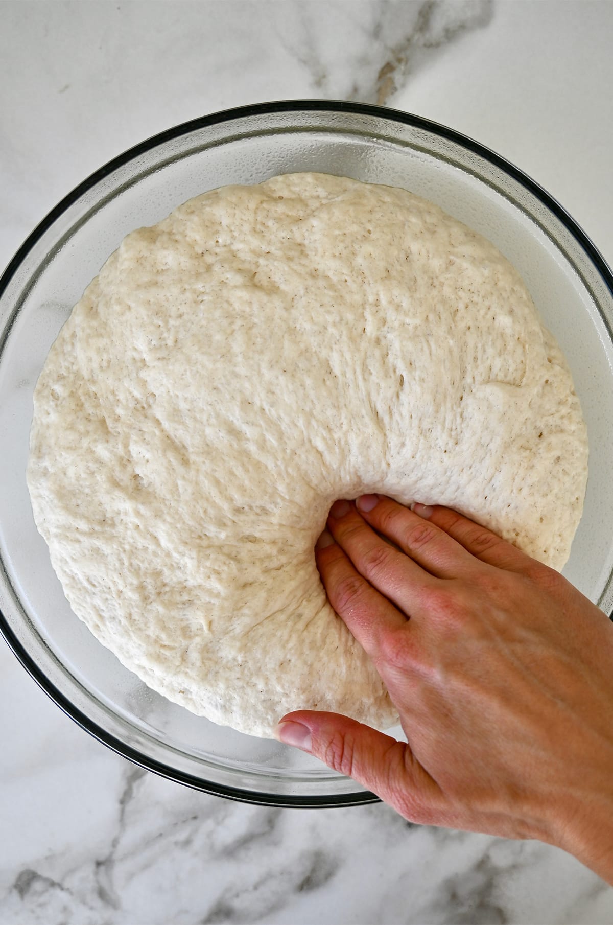 A hand presses into proofed pretzel dough in a glass bowl.