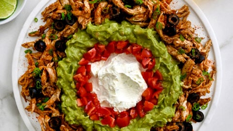 A top-down view of a white platter containing Rotisserie Chicken Taco Dip surrounded by limes and bowls of chips