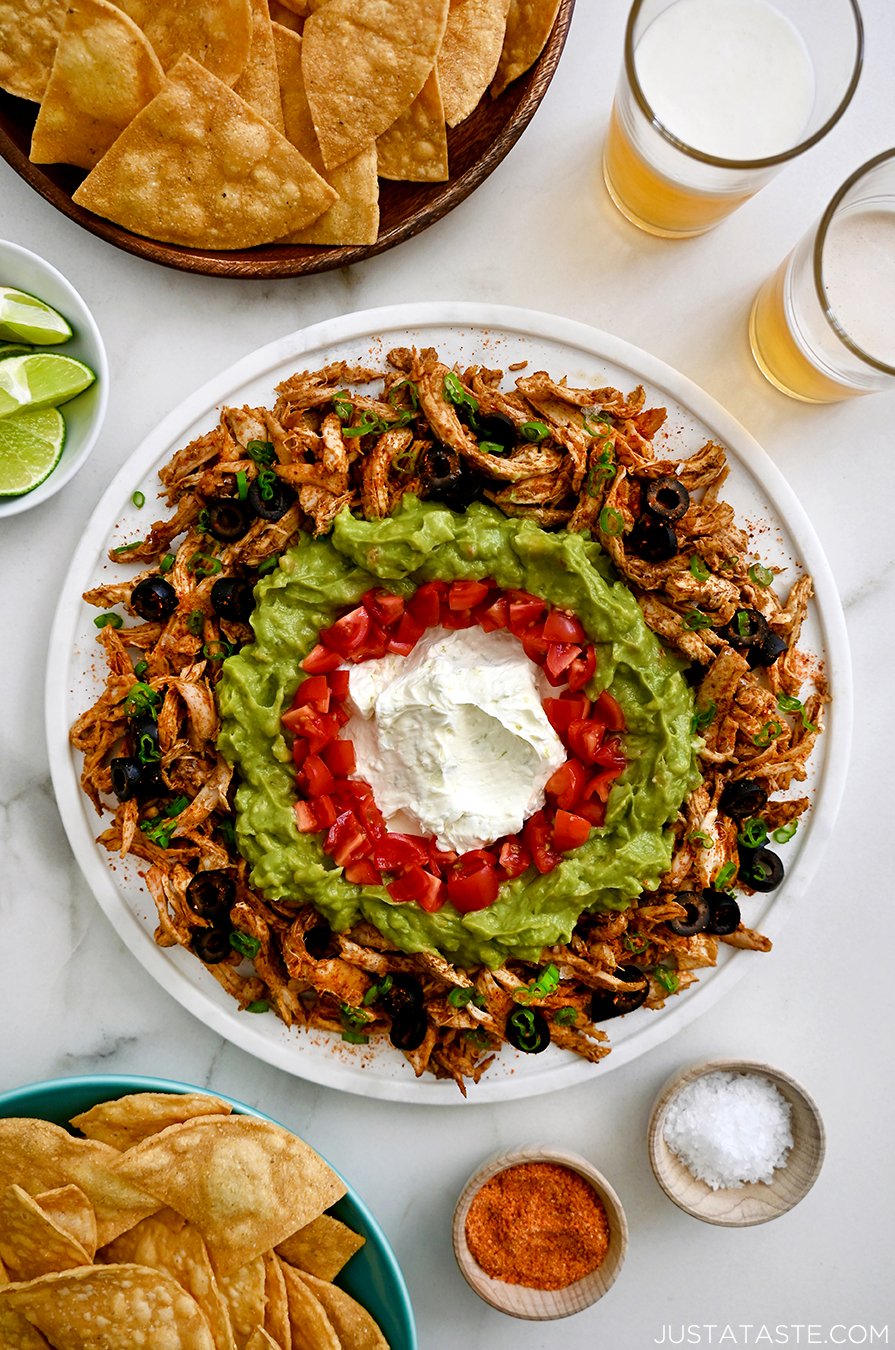 A top-down view of a white platter containing Rotisserie Chicken Taco Dip surrounded by limes and bowls of chips