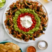 A white platter with taco dip surrounded by bowls of chips, seasonings, and two glasses of beer.