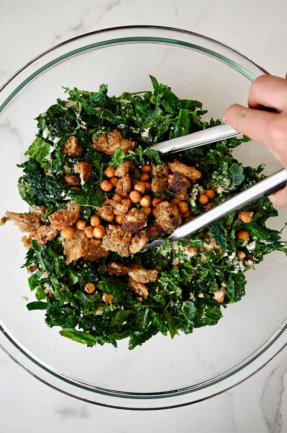 Using tongs to toss Caesar dressing with shredded kale, crispy chickpeas and croutons.