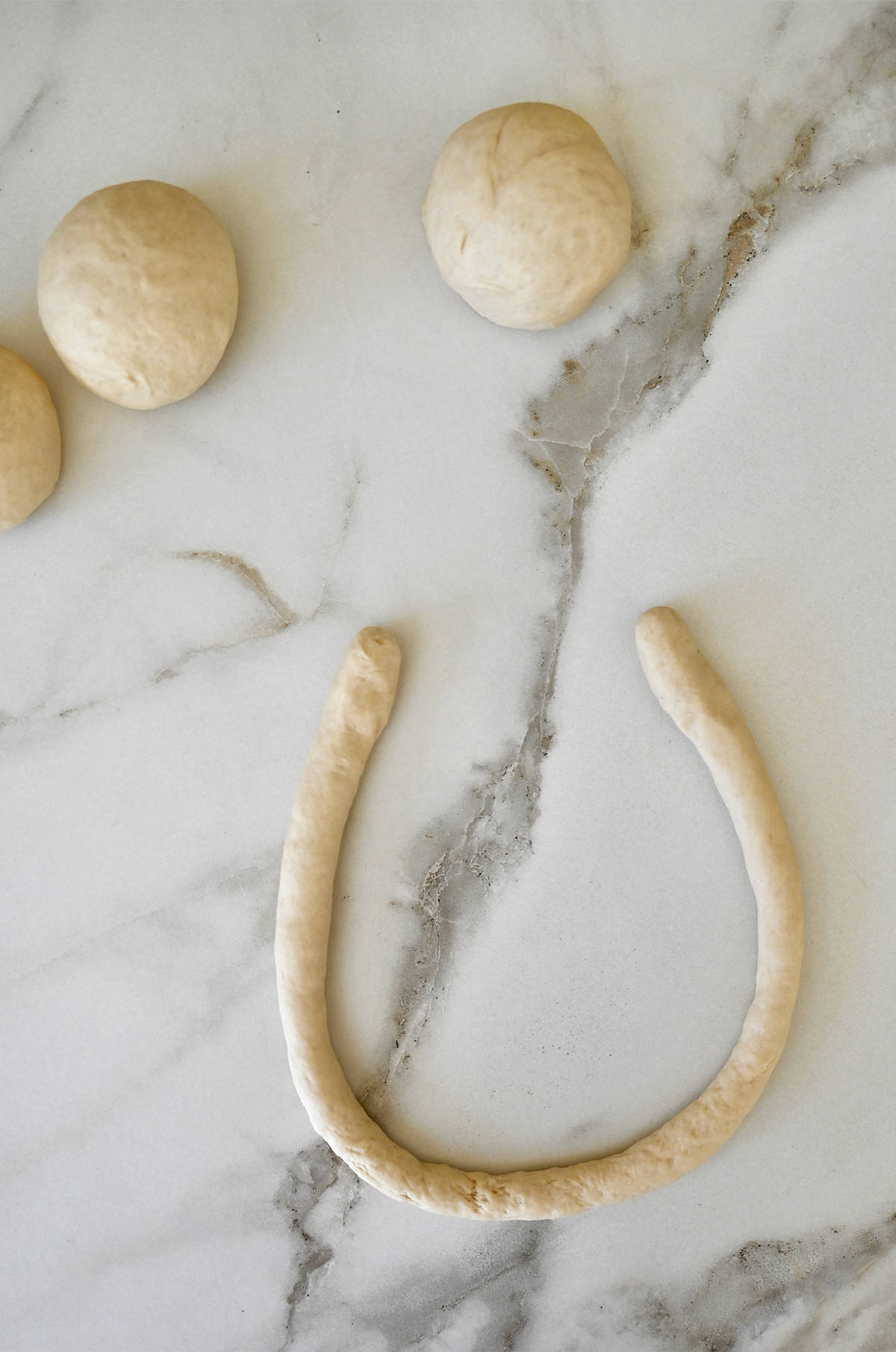 A long rope of dough in a "u" shape on a marble surface next to three balls of dough.