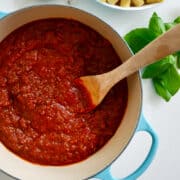 A large stock pot containing Secret Ingredient Bolognese Sauce and a wooden spoon next to a large bowl containing rigatoni noodles.