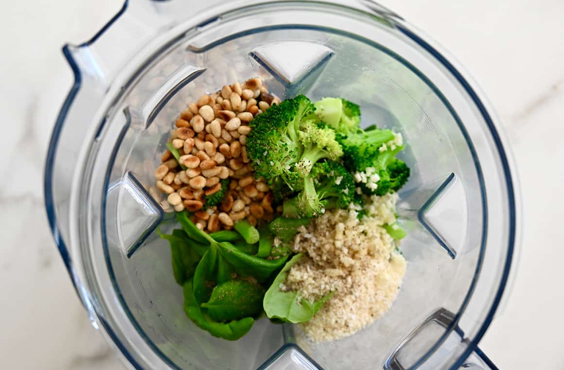 A top-down view of a blender containing basil, pine nuts, broccoli, Parmesan and garlic