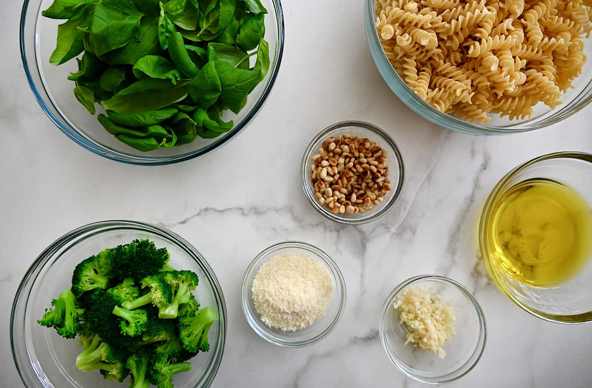 Various sizes of bowls containing basil, uncooked pasta, olive oil, pine nuts, garlic, parmesan, and broccoli