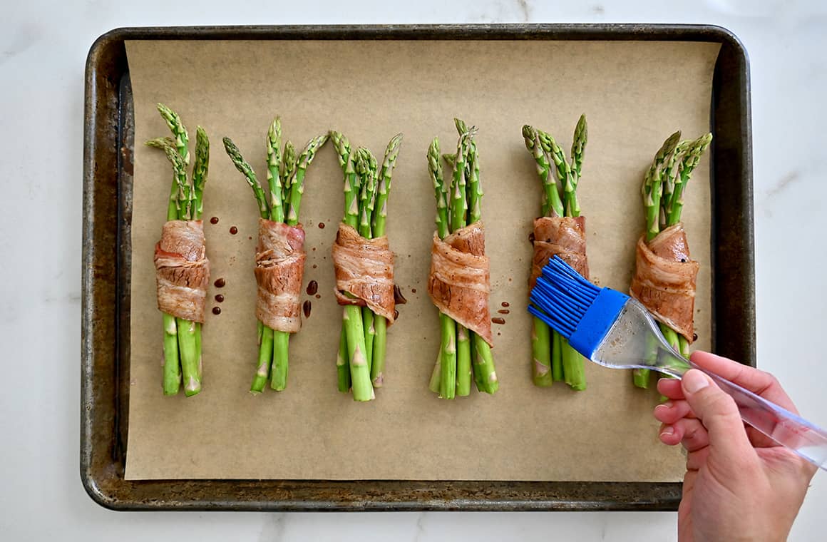A hand with a food brush, brushes balsamic vinegar atop asparagus spears wrapped with bacon on a parchment paper-lined baking sheet