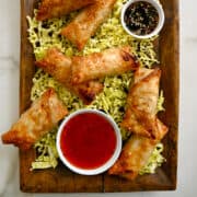 A top-down view of air fryer spring rolls atop a bed of shredded cabbage on a serving platter with a bowl filled with sweet chili sauce and another filled with soy dipping sauce.