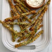 A top-down view of Panko-crusted Asparagus Fries on a white, Le Creuset serving platter with a small bowl containing roasted garlic aioli.