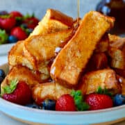A pile of baked French toast sticks on a plate with fresh berries that are all being drizzled with maple syrup.