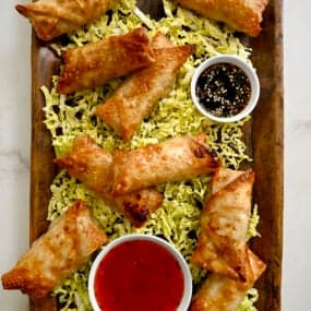 A top-down view of a wood serving plate with Air Fryer Spring Rolls on a bed of shredded cabbage next to small bowls containing sweet chili sauce and soy dipping sauce