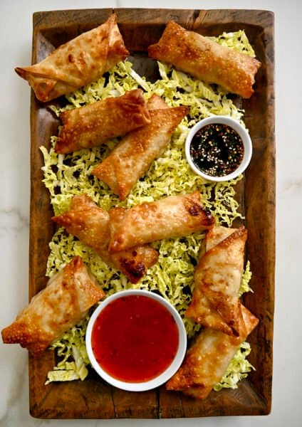 A top-down view of a wood serving plate with Air Fryer Spring Rolls on a bed of shredded cabbage next to small bowls containing sweet chili sauce and soy dipping sauce