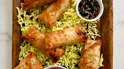A top-down view of a wood serving plate with Air Fryer Spring Rolls on a bed of shredded cabbage next to small bowls containing sweet chili sauce and soy dipping sauce