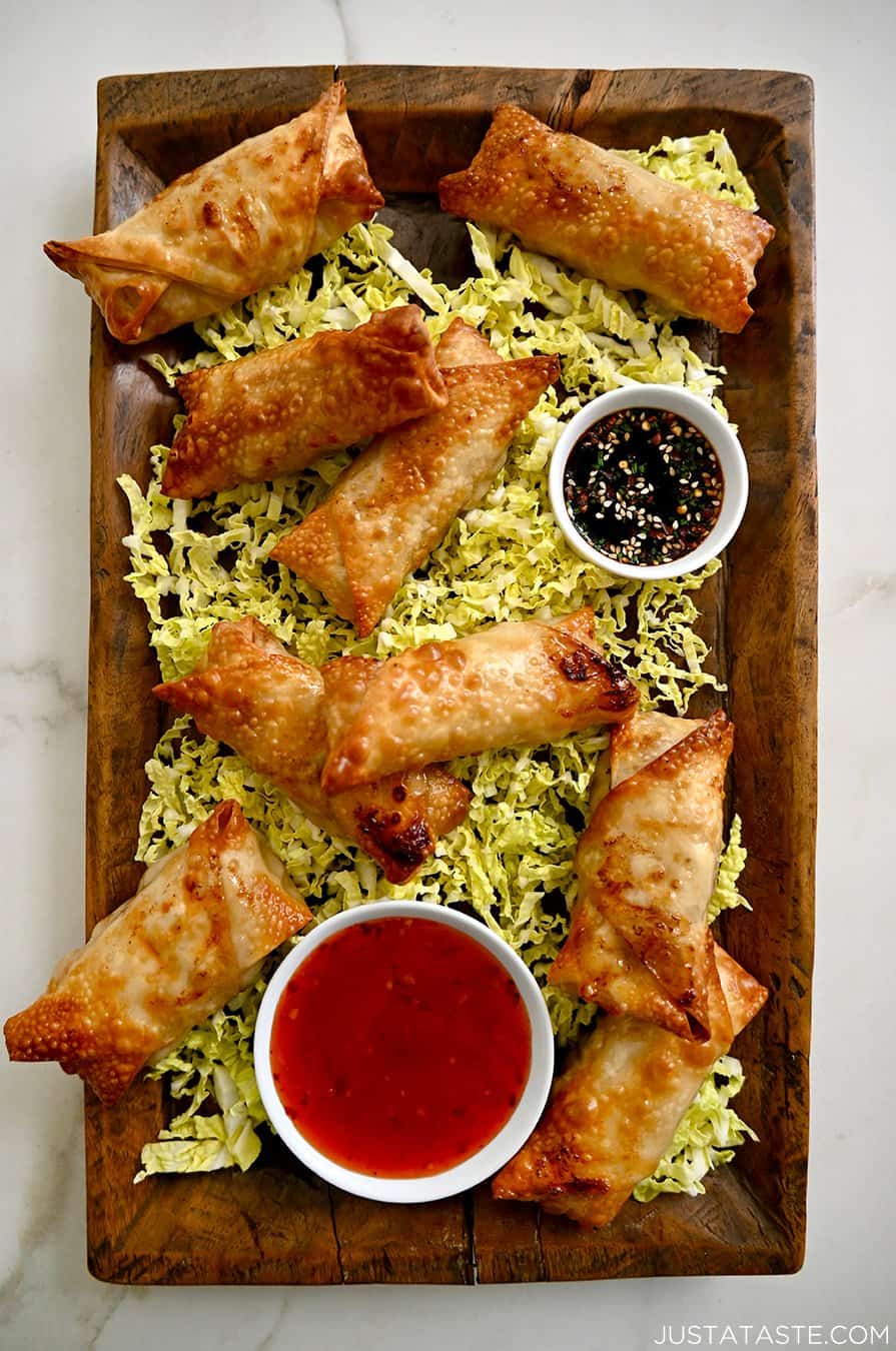 A top-down view of a wood serving plate with Air Fryer Spring Rolls on a bed of shredded cabbage next to small bowls containing sweet chili sauce and soy dipping sauce