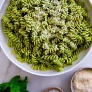 A large white bowl containing Broccoli Pesto Pasta topped with parmesan cheese next to a wooden spoon, basil leaves and a small bowl filled with parmesan.