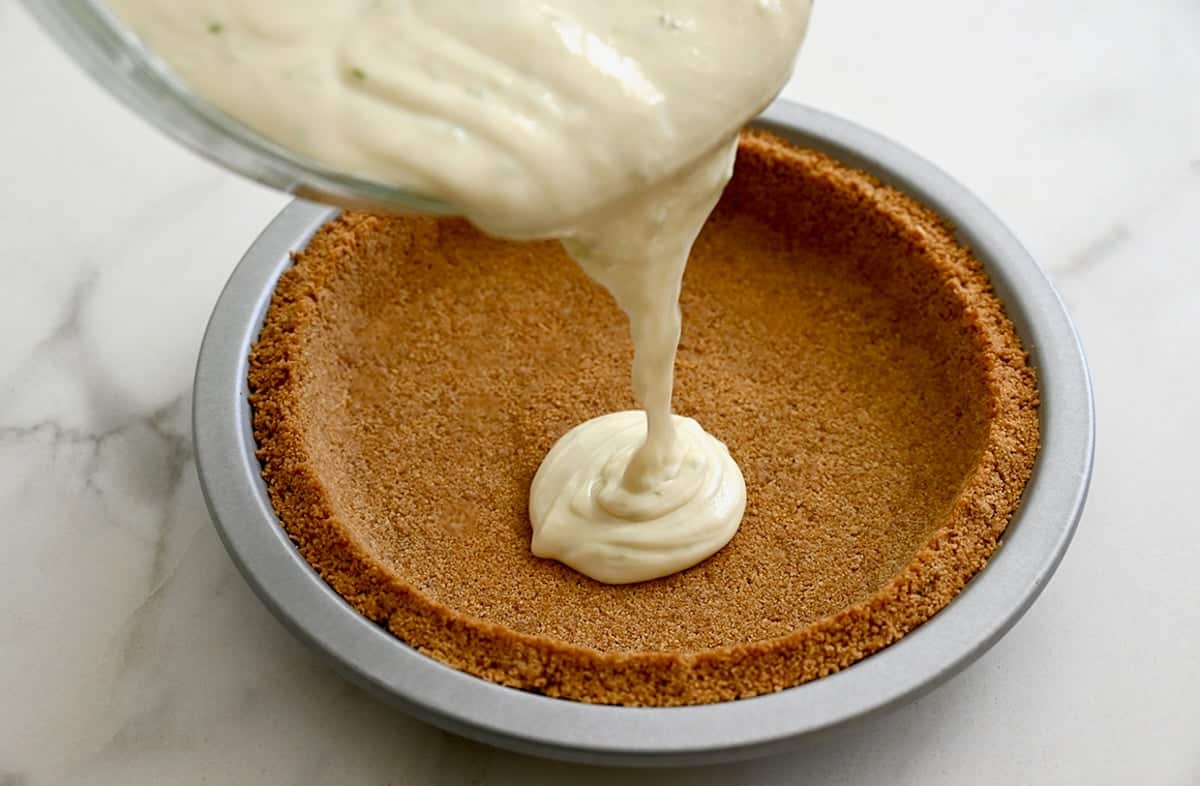 Key lime pie filling is being poured from a bowl into a graham cracker crust.