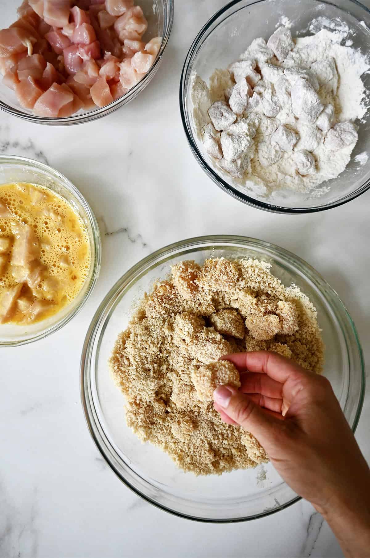 Four glass bowls containing chicken pieces, flour, egg wash and Panko breadcrumbs. This is the process for breading the chicken before baking.