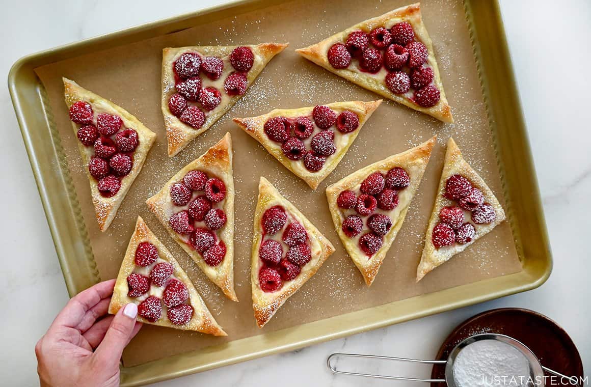 A hand reaches for a cream cheese danish with raspberries that's dusted with confectioners' sugar