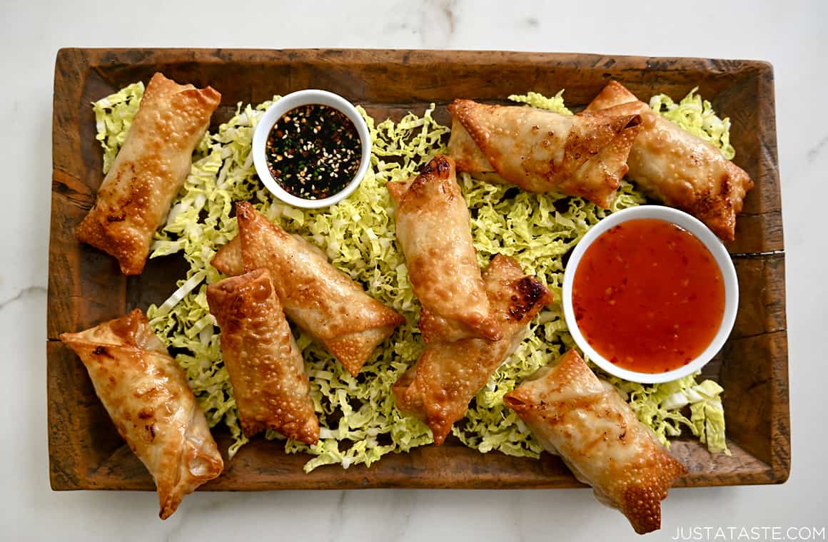 A wood serving plate with Air Fryer Spring Rolls and small bowls containing sweet chili sauce and soy dipping sauce atop a bed of shredded cabbage 